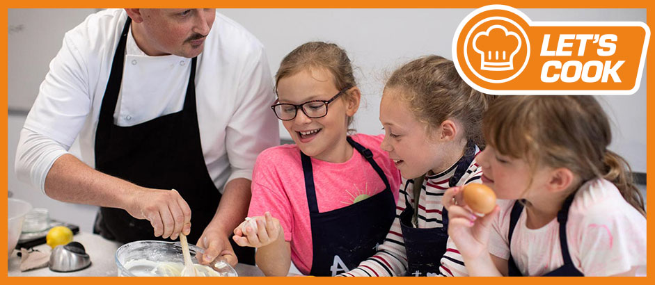Three children cooking at a cooking camp, look on as a chef engages them in fun